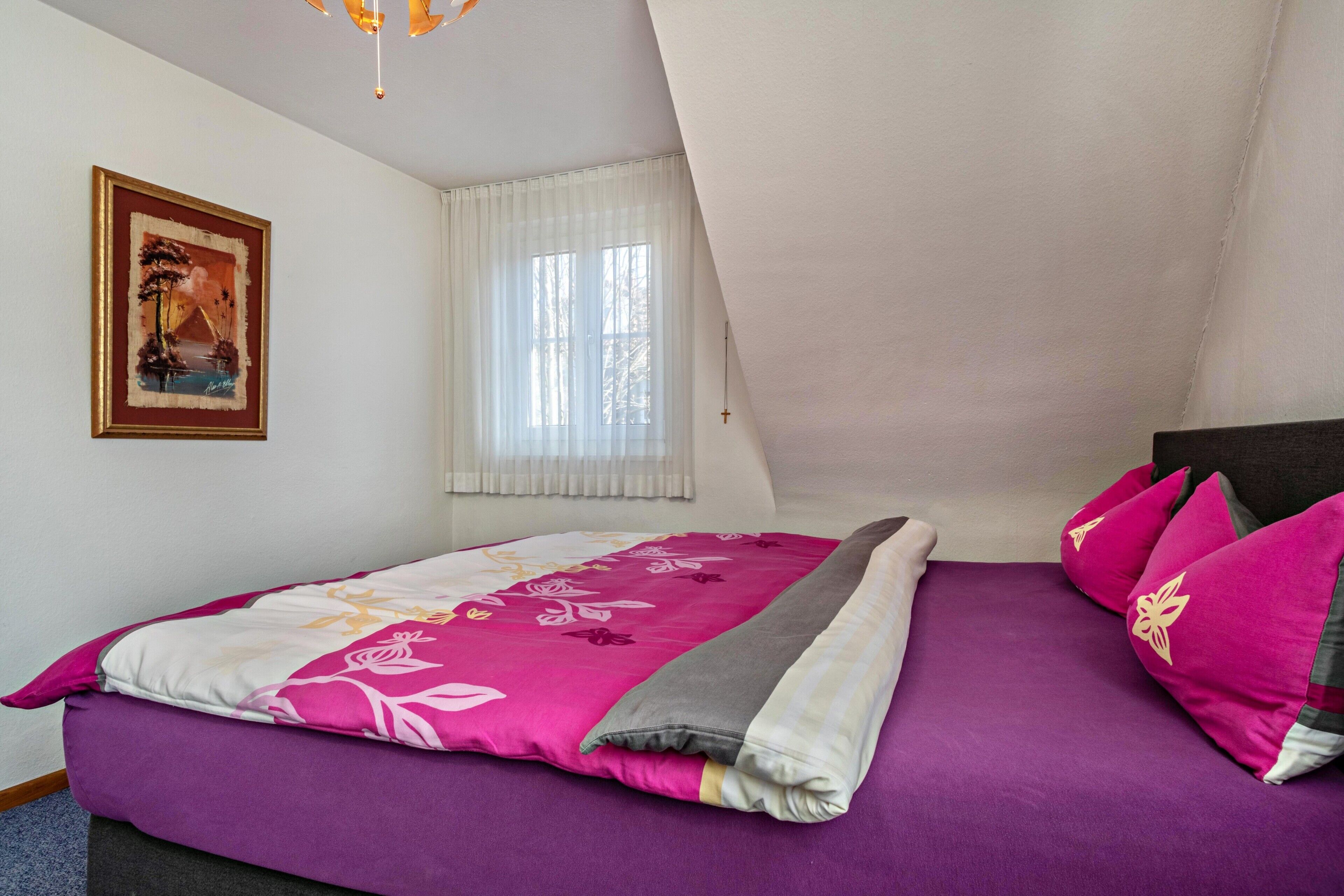 Une chambre à coucher avec un lit, une fenêtre avec des rideaux, un tableau au mur et une croix accrochée au mur.
