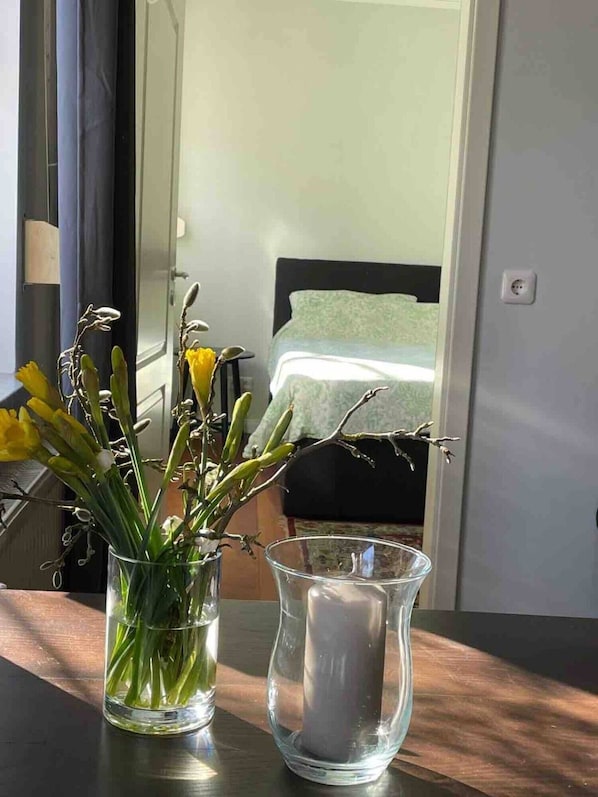 A room with a bed, a window, a vase with flowers, and a candle on a table.