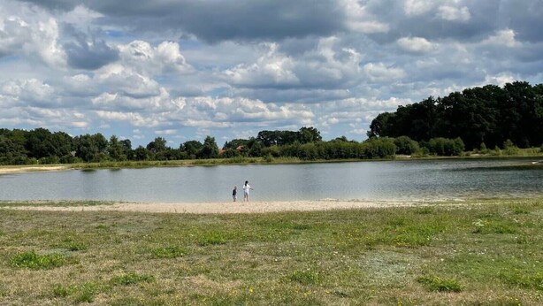 En sjö med en sandstrand, omgiven av träd och en molnig himmel.