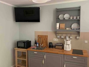 A compact kitchen with a microwave, toaster, coffee maker, and a wooden shelf with dishes.
