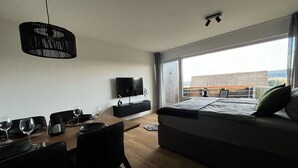 A modern hotel room with a large bed, a dining area with a table and chairs, a TV mounted on the wall, and a balcony with a view.