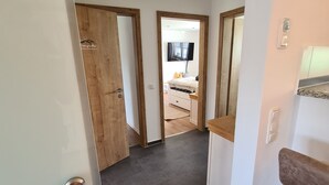 Un pasillo con una puerta de madera, pared blanca, techo con iluminación empotrada y una vista de un dormitorio con cama y televisor.