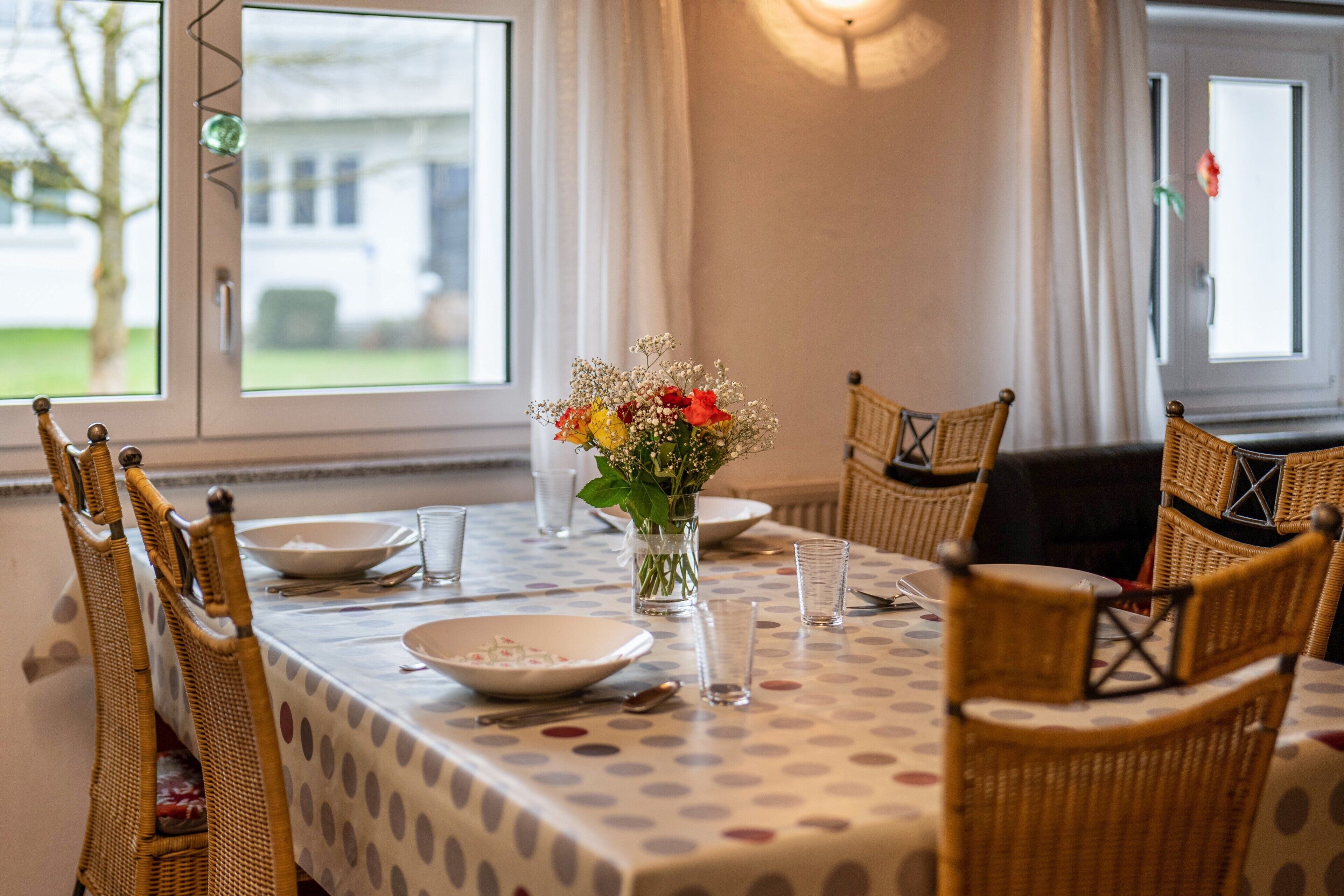 A dining table with a polka dot tablecloth, surrounded by wicker chairs, and a vase with flowers.