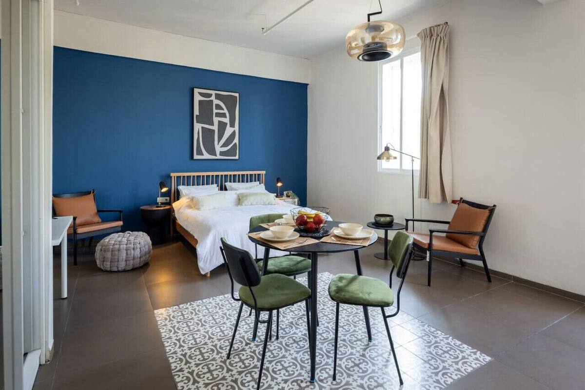A modern bedroom with a blue accent wall, a bed with white linens, a dining table with chairs, and a window with curtains.