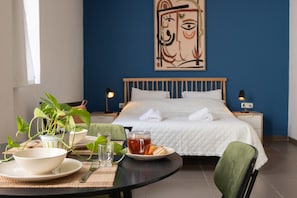 Une table de petit-déjeuner bien rangée, avec un bol, une assiette et un verre, une plante en pot et une tasse de thé. En arrière-plan, on aperçoit un lit avec une literie blanche et un mur bleu orné d’un tableau.
