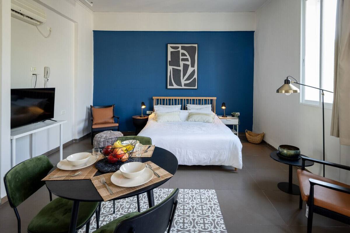 A modern bedroom with a large bed, a dining area with chairs and a table, a television, and a blue accent wall.