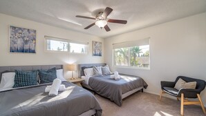 A hotel room with two beds, a ceiling fan, artwork on the walls, and a window with a view of palm trees.