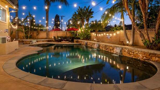 A swimming pool with a waterfall feature, surrounded by palm trees and string lights.