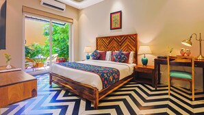 A bedroom with a bed, bedside tables, a desk, and a chair. There is a window with a view of greenery outside. - Ananyamaya Nature Lodge (Sawai Madhopur)