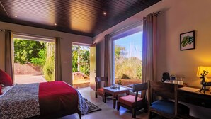 A bedroom with a bed, a desk, and chairs, large windows, and a view of the outdoors. - Ananyamaya Nature Lodge (Sawai Madhopur)