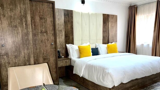 A hotel room with a large bed, a wooden headboard, a round glass table with a glass vase, and a view of the outdoors. - UrbaN RootZ  (Abuja)