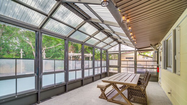 A covered patio with a table and chairs, large windows, and a view of trees. - Poolside Paradise! Central Merritt Island Home (Merritt Island)