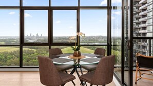 A modern room with a large window, a round glass table with four chairs, and a view of a cityscape and greenery. - Chic 1-Bed with Pool by River, Ferry & Amenities (Wentworth Point)