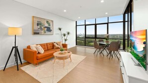 A modern living room with a large window, a sofa, a coffee table, and a painting. - Chic 1-Bed with Pool by River, Ferry & Amenities (Wentworth Point)