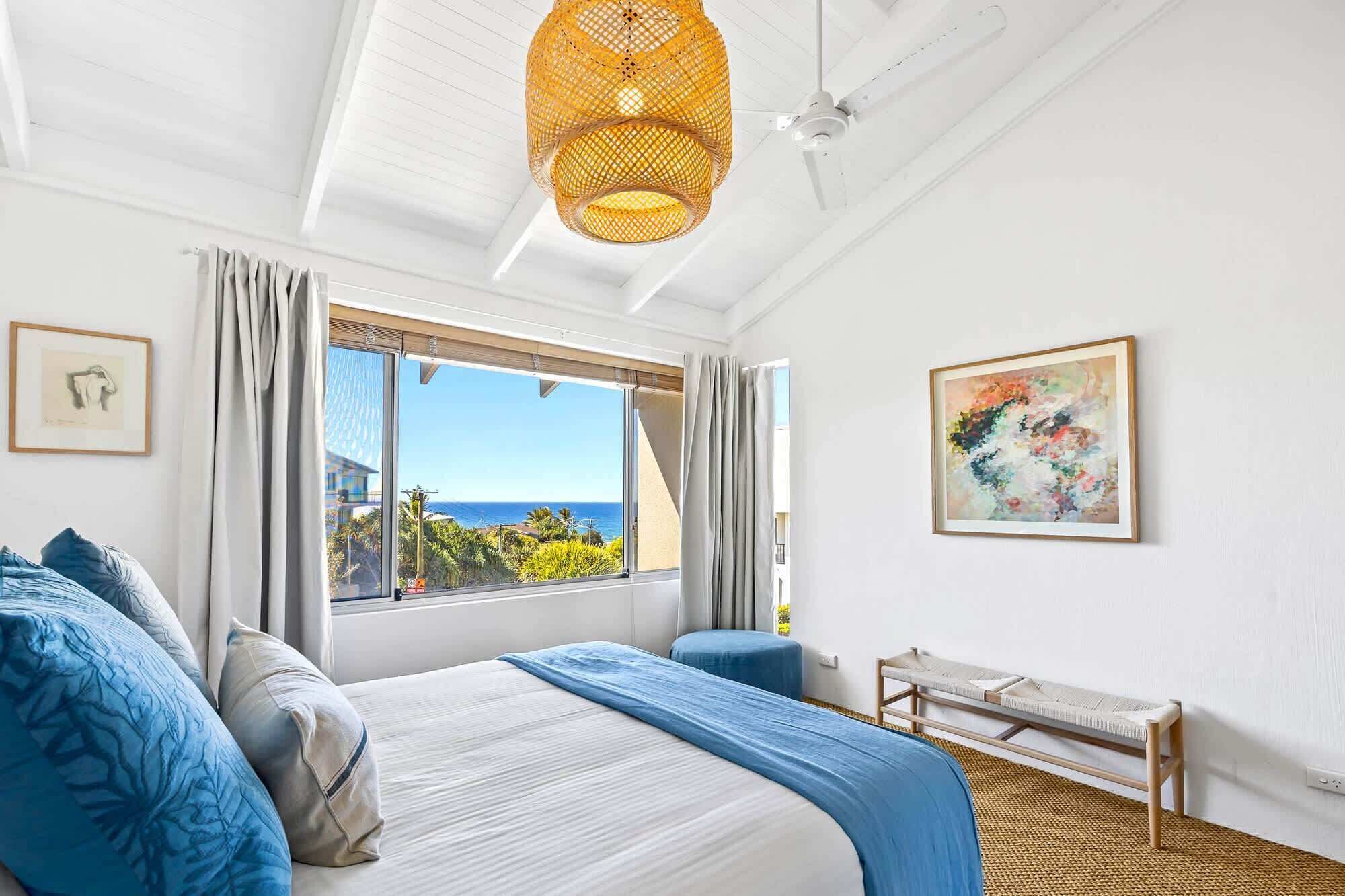 A hotel room with a large bed, a view of the ocean, a ceiling fan, and a painting on the wall.