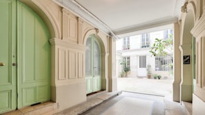 A hallway with a green door, a tiled floor, and a view of a courtyard with potted plants and a building in the background.