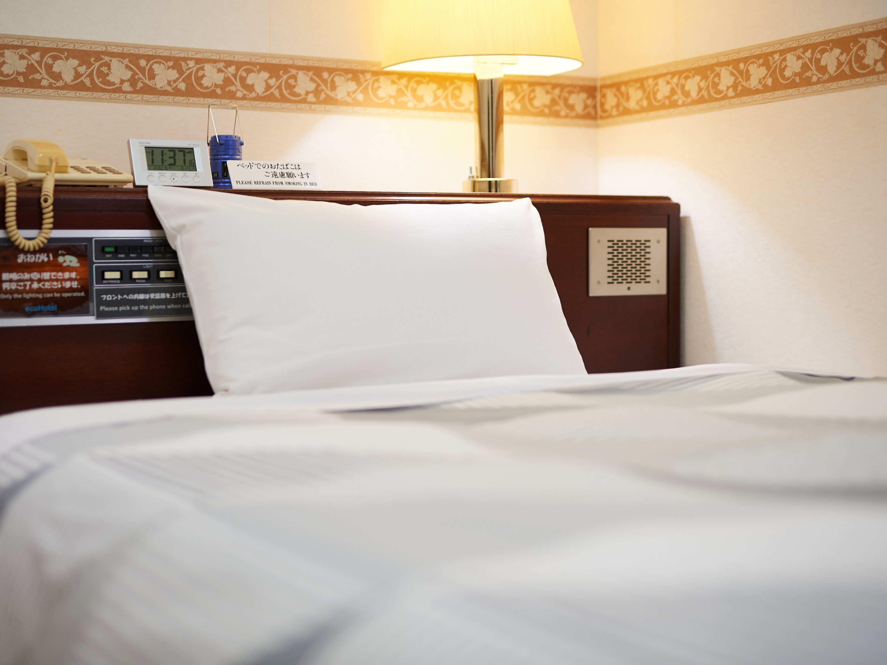 A hotel room with a bed, a bedside table with a lamp, a clock, and a phone.