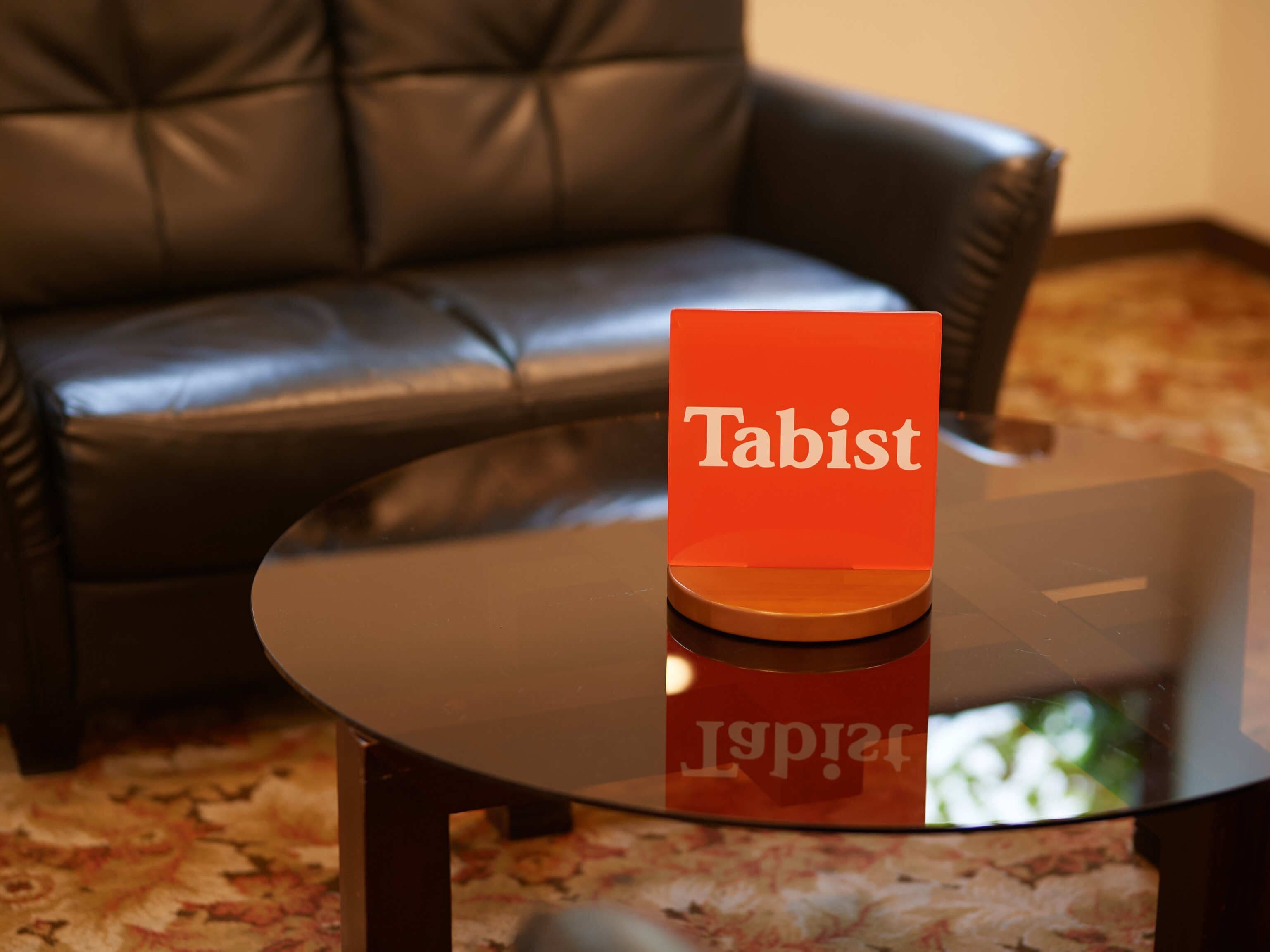 A glass table with a red sign labeled "Tabist" in a room with a black leather couch.