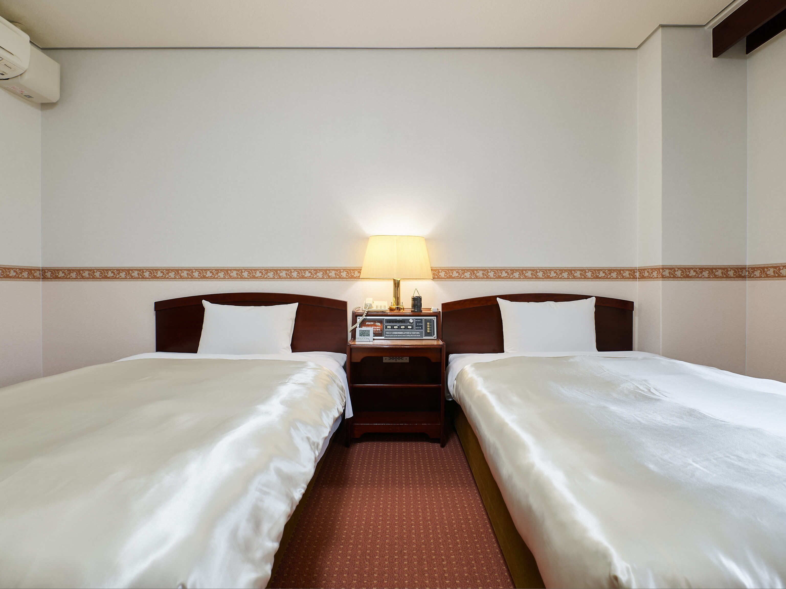 Habitación de hotel con dos camas individuales, una mesita de noche con lámpara y aire acondicionado instalado en la pared.