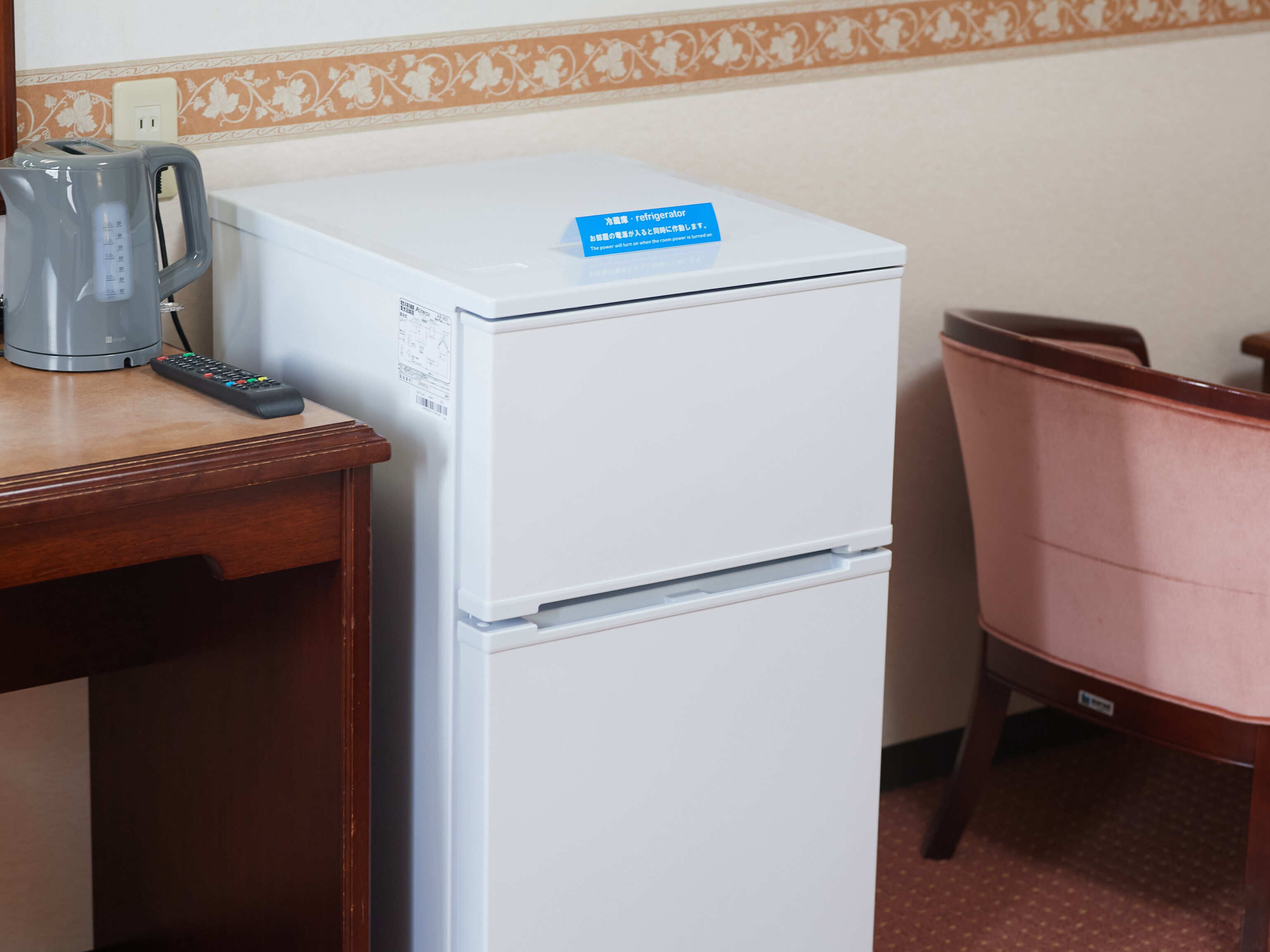 Ein weißer Kühlschrank mit blauer Beschriftung, eine Fernbedienung und ein Wasserkocher auf einem Holztisch.