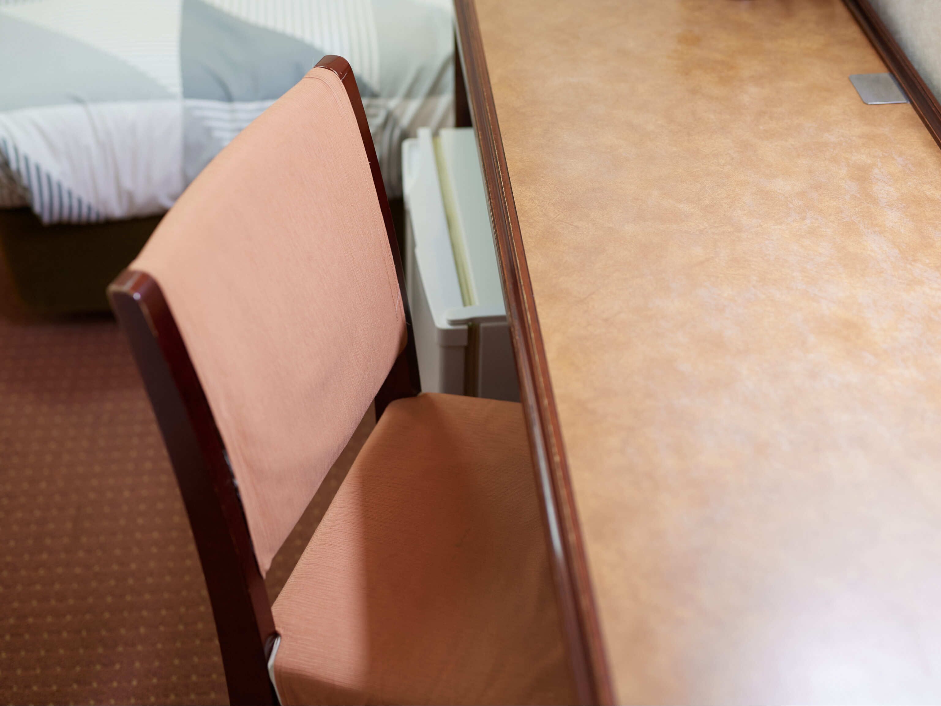 Una mesa de madera con una placa metálica, una silla con cojín marrón y una cama con ropa de cama a rayas al fondo.