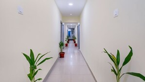 A long, narrow hallway with white walls and a tiled floor, flanked by potted plants and doors leading to other rooms. - iROOMZ KSR Luxury Rooms (Hyderabad)