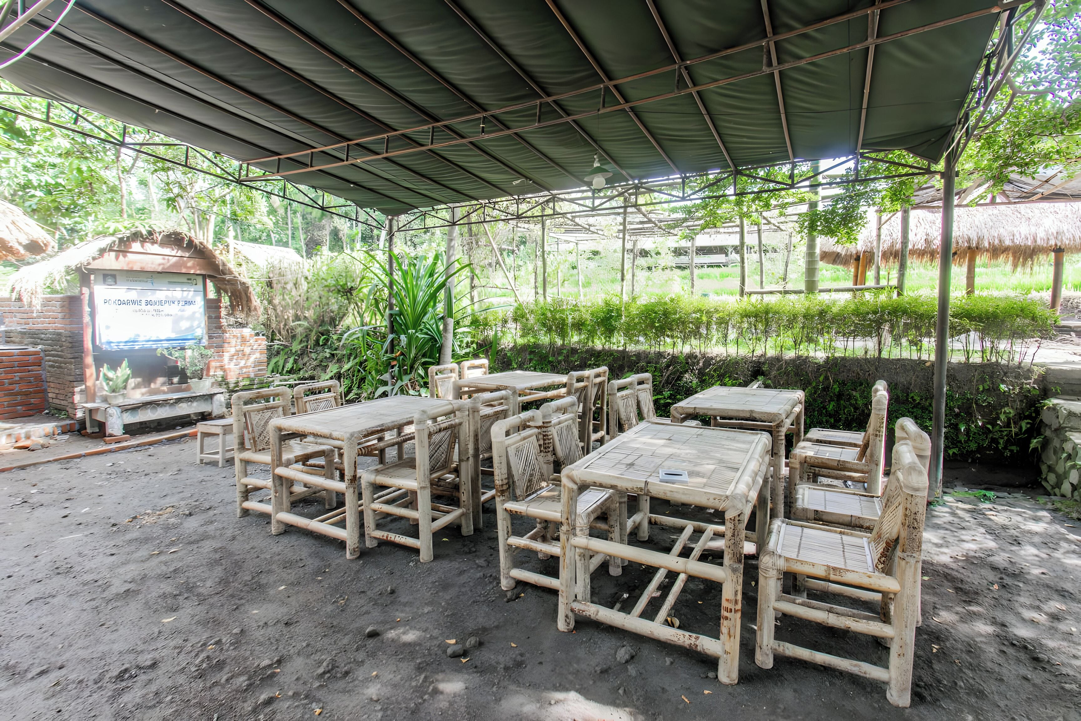 A covered outdoor seating area with tables and chairs, surrounded by greenery and a brick structure.