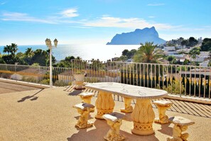 A terrace with a table and chairs, overlooking a coastal view with a mountain in the background.
