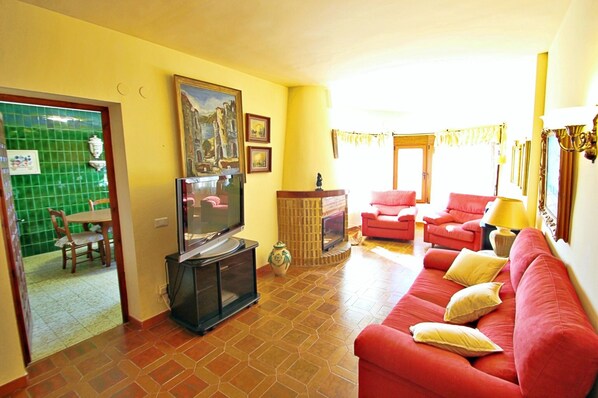 A living room with a red sofa, a TV, and a fireplace. There is a dining area with a table and chairs visible through an open door.