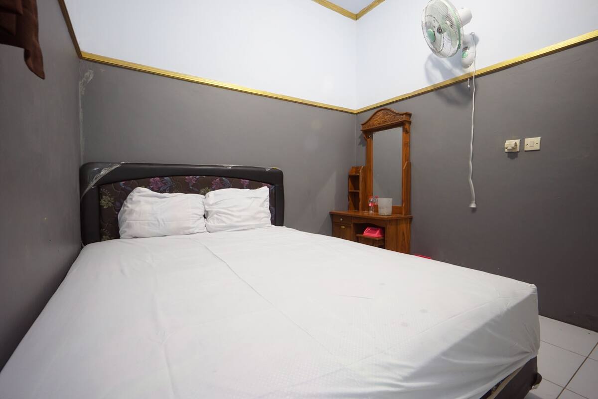 Une chambre à coucher comprenant un lit, une table de chevet, un miroir et un ventilateur fixé au mur.