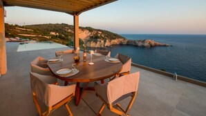 A dining area with a wooden table and chairs, overlooking a coastal landscape with cliffs and the sea. - Seawhisper Villa in Paxi (Paxos)