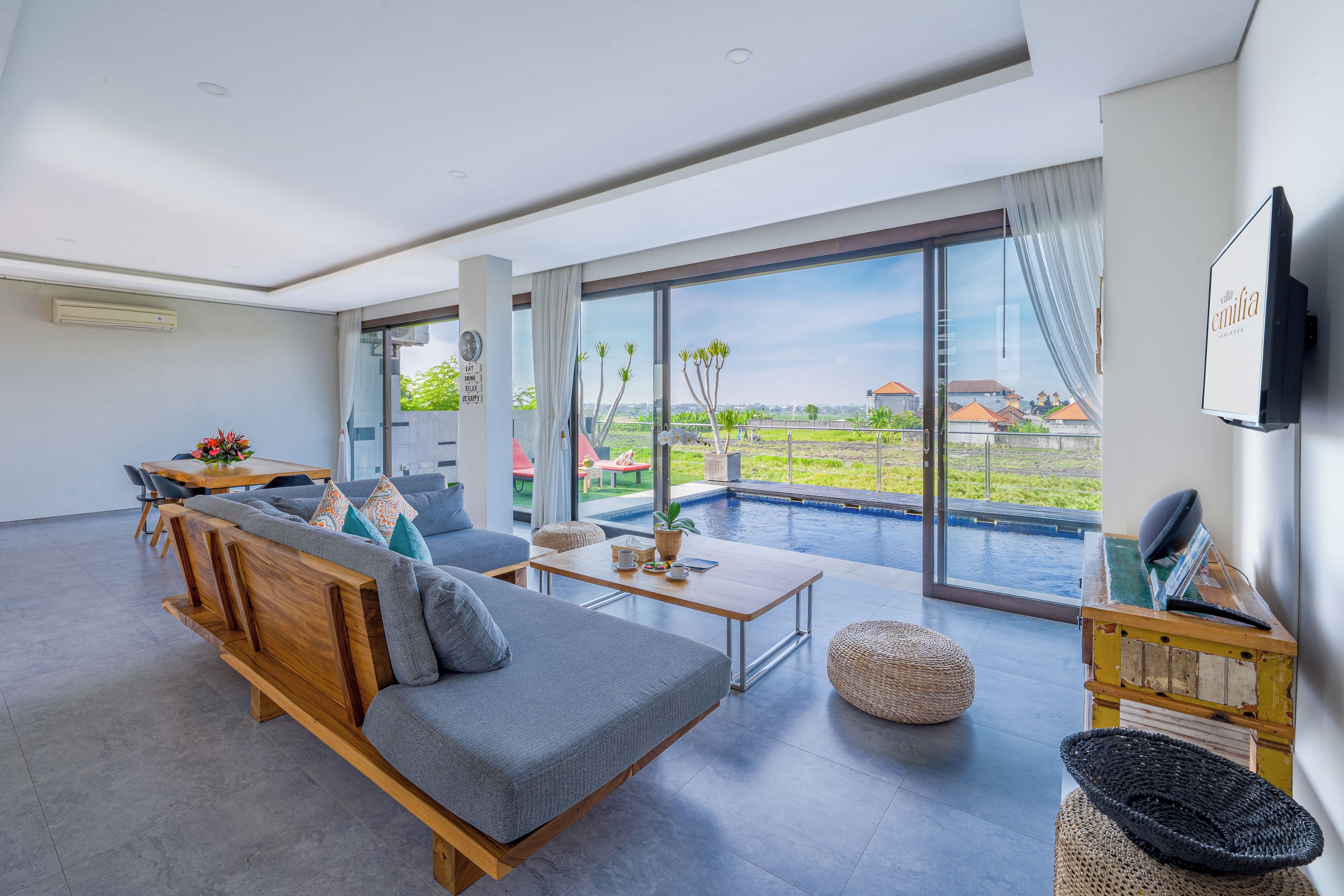 Una sala de estar moderna con un sofá, una mesa de centro y un televisor. Hay un ventanal con vistas a una piscina y casas.