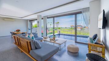 Una sala de estar moderna con un sofá, una mesa de centro y un televisor. Hay un ventanal con vistas a una piscina y casas.