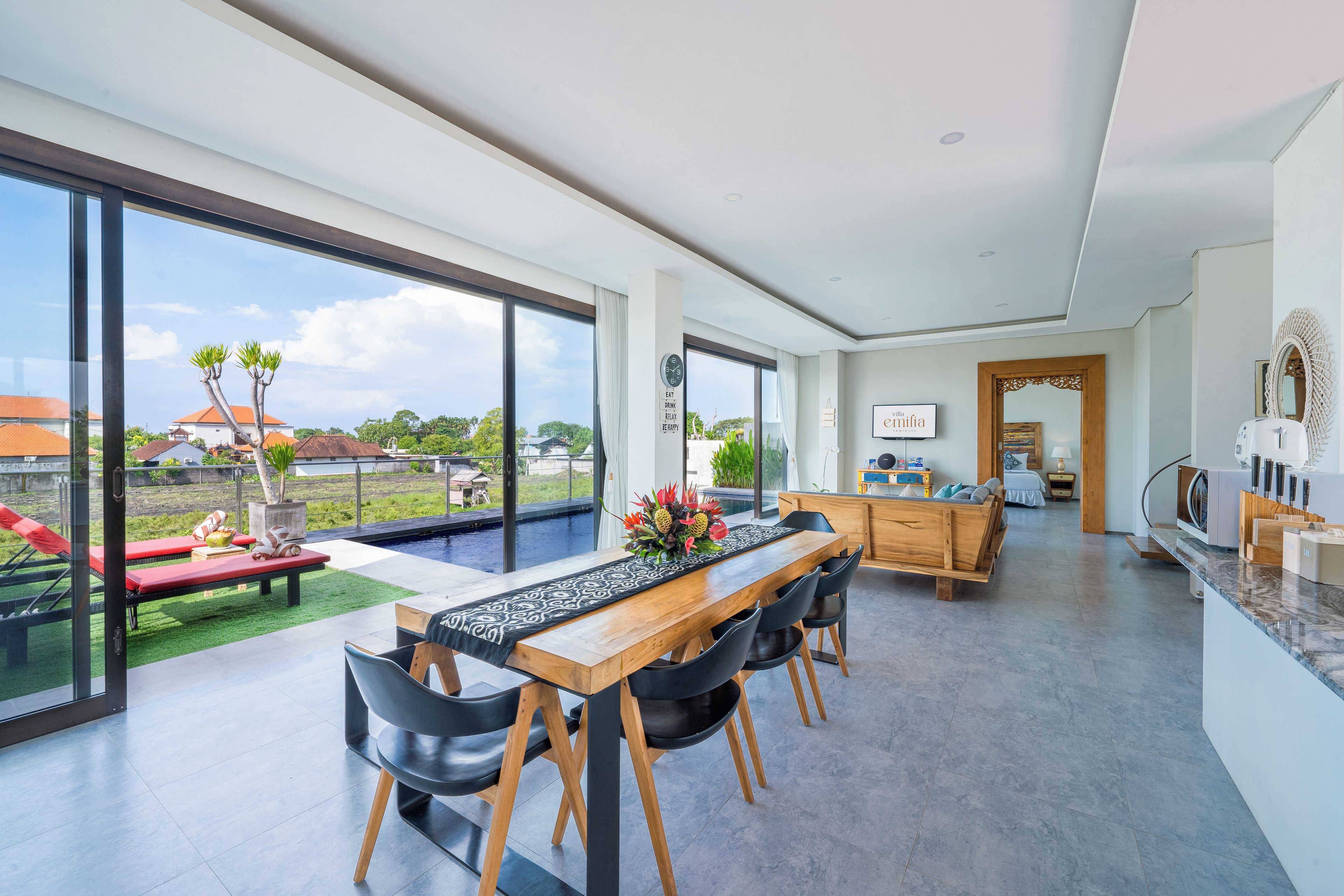 A spacious living area with a dining table, chairs, and a view of a swimming pool and greenery.