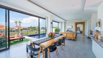 A spacious living area with a dining table, chairs, and a view of a swimming pool and greenery.