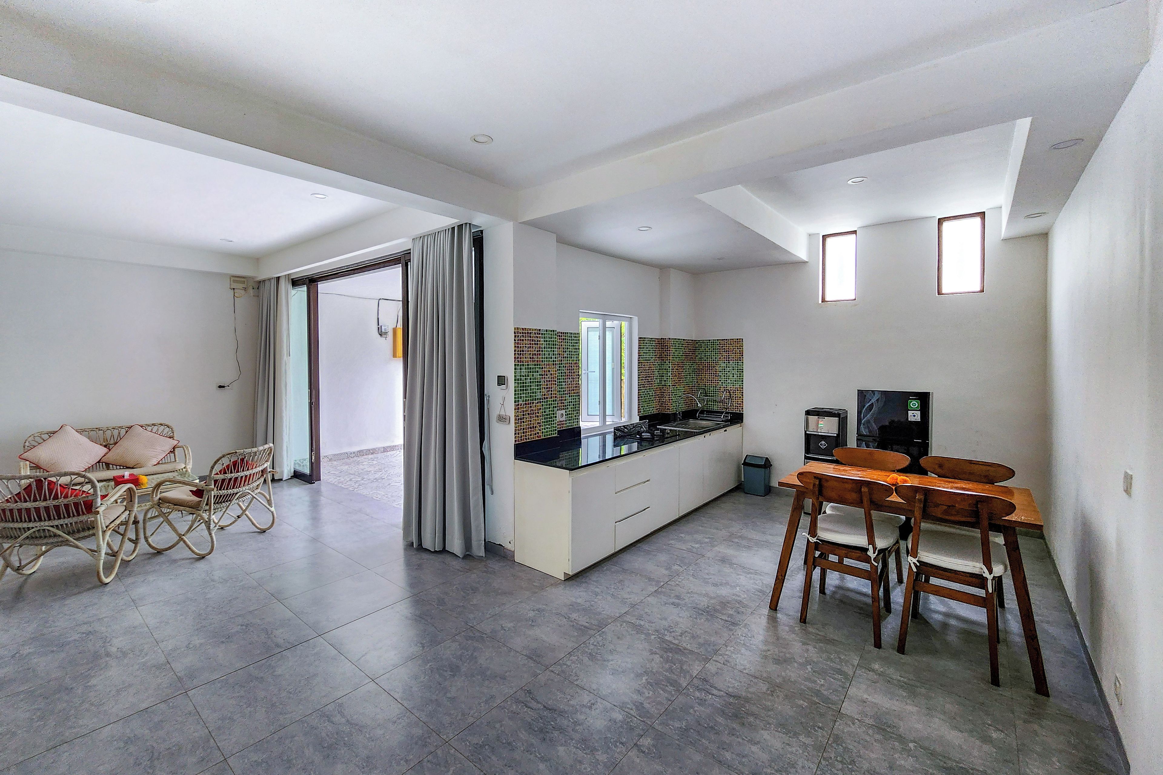 A modern kitchen with a black countertop, white cabinets, and a small window. There is a dining area with a wooden table and chairs, and a wicker sofa with cushions.