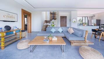 Una sala de estar moderna con una mesa de centro de madera, un sofá gris con cojines azules, una escalera y una cocina visible a través de una puerta abierta.