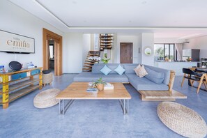 A modern living room with a wooden coffee table, a grey sofa with blue cushions, a staircase, and a kitchen area visible through an open door. - Villa Emilia Seminyak by Nagisa Bali (Kerobokan)