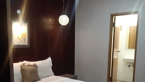 A neatly made bed with white linens and a folded towel, a wooden headboard, a wall-mounted light fixture, and a door leading to a bathroom. - Hotel Djagalan Raya  (Surabaya)