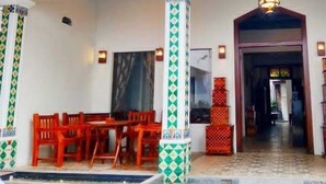 A covered outdoor seating area with wooden chairs and a table, a tiled floor, and a decorative column with a geometric pattern. - Hotel Djagalan Raya  (Surabaya)