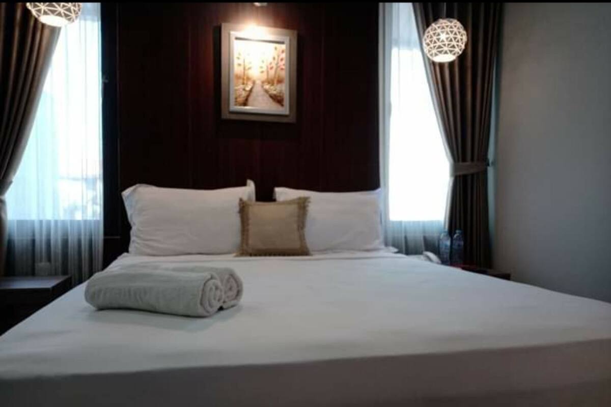 Un lit bien fait, avec du linge de lit blanc et une serviette enroulée, une œuvre d’art encadrée au-dessus du lit, et une fenêtre avec des rideaux de chaque côté.