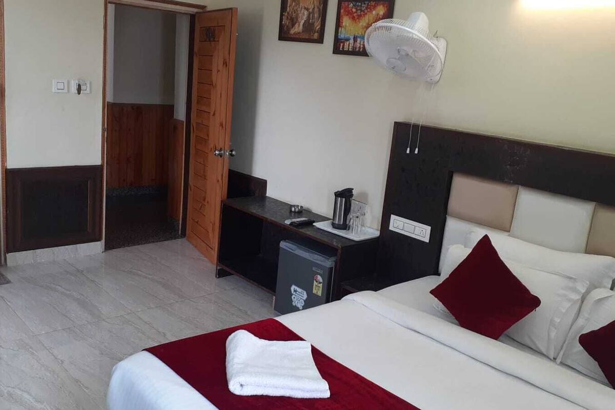 Una habitación de hotel con una cama, un escritorio, un refrigerador pequeño, un ventilador de pared y una puerta que da a otra habitación.