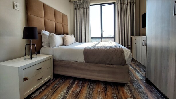 A modern hotel room with a large bed, a bedside table with a lamp, a wardrobe, and a large window with curtains. - Zwelakho Luxury  furnished apartments (Sandton)