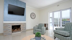 A living room with a fireplace, a flat-screen TV, a clock, striped armchairs, and a round coffee table with a plant. - Zwelakho Luxury  furnished apartments (Sandton)