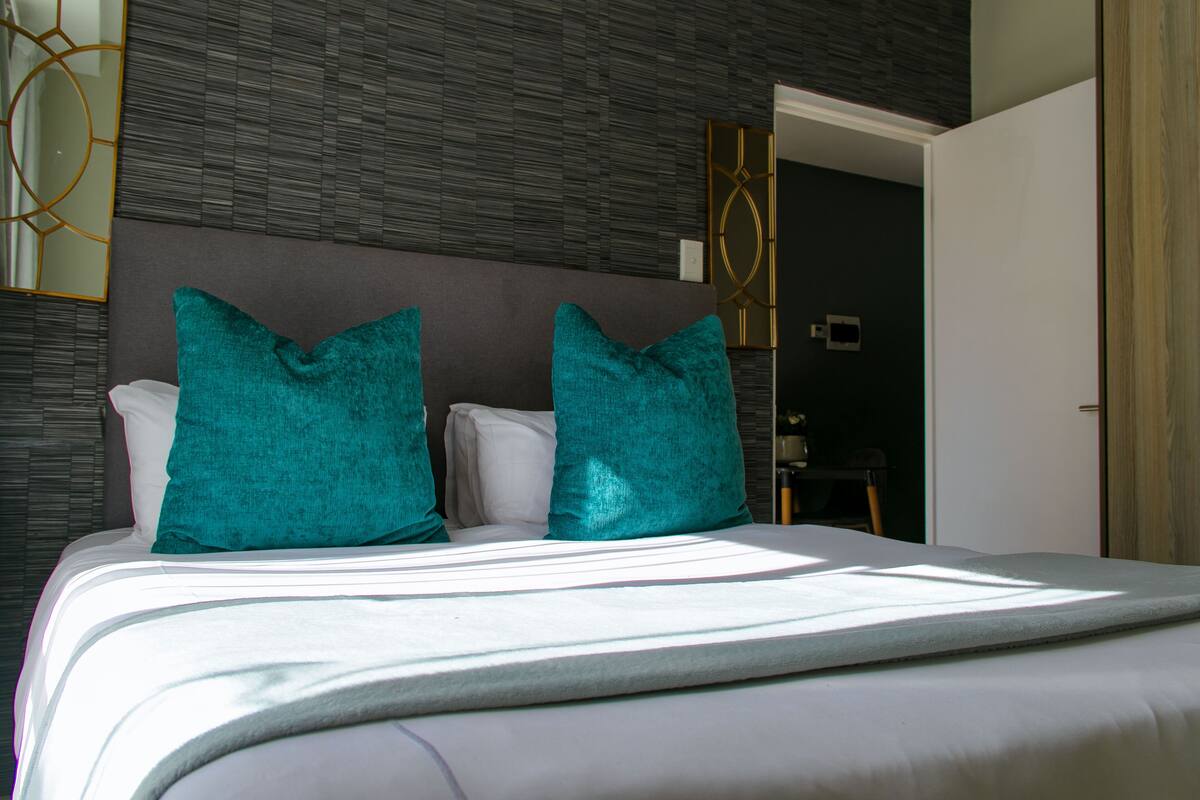 Ein ordentlich bezogenes Bett mit weißen Leinen und türkisfarbenen Kissen, eine moderne Wand mit einem geometrischen Spiegel und eine teilweise geöffnete Tür zu einem anderen Raum.