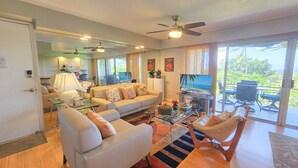 A spacious living room with a sofa, armchairs, a coffee table, and a TV. There is a sliding glass door leading to an outdoor patio area. - Ocean View Kona Condo | Relax, Unwind & Enjoy Island Life (Kailua-Kona)