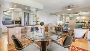A spacious living area with a glass dining table, comfortable seating, a kitchen area, and a ceiling fan. - Ocean View Kona Condo | Relax, Unwind & Enjoy Island Life (Kailua-Kona)