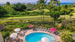 A pool area with lounge chairs and umbrellas, surrounded by lush greenery and palm trees, with a view of the ocean and golf course in the background. - Ocean View Kona Condo | Relax, Unwind & Enjoy Island Life (Kailua-Kona)