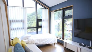 Un dormitorio moderno con cama, sofá, televisor y ventanas amplias que ofrecen vistas al exterior.