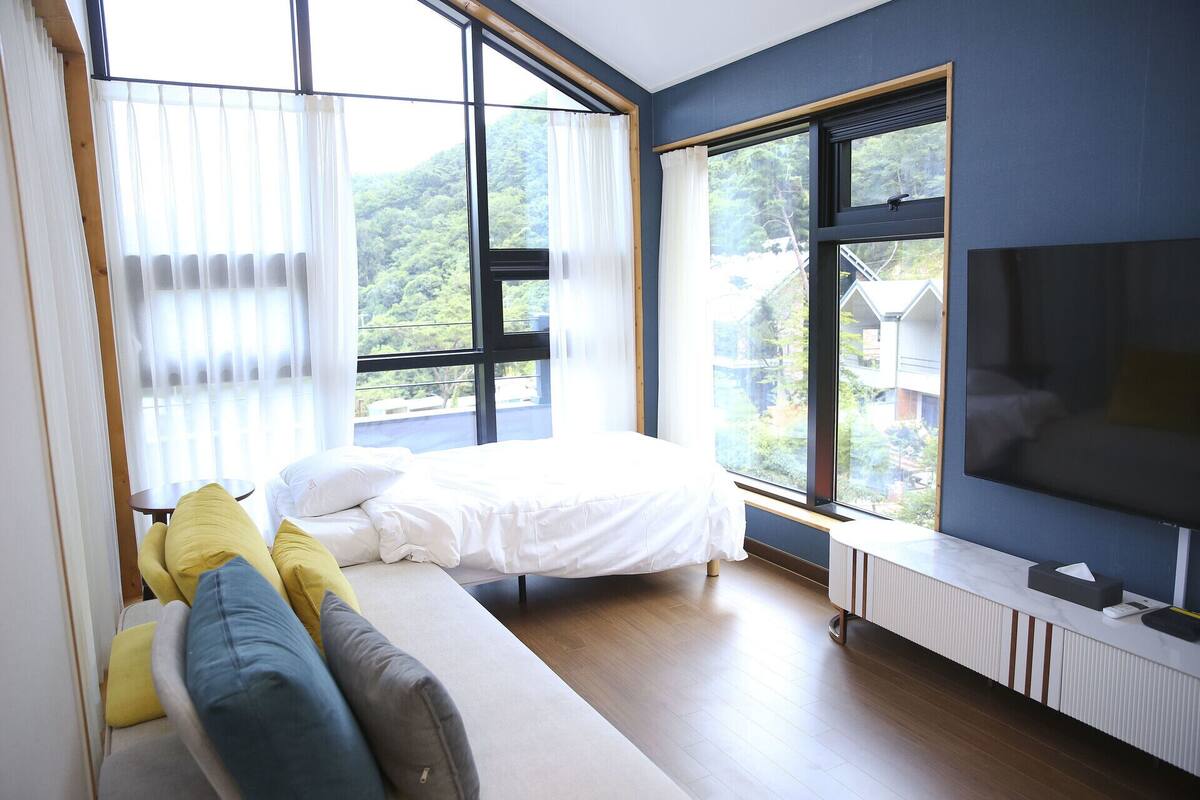 Un dormitorio moderno con cama, sofá, televisor y ventanas amplias que ofrecen vistas al exterior.